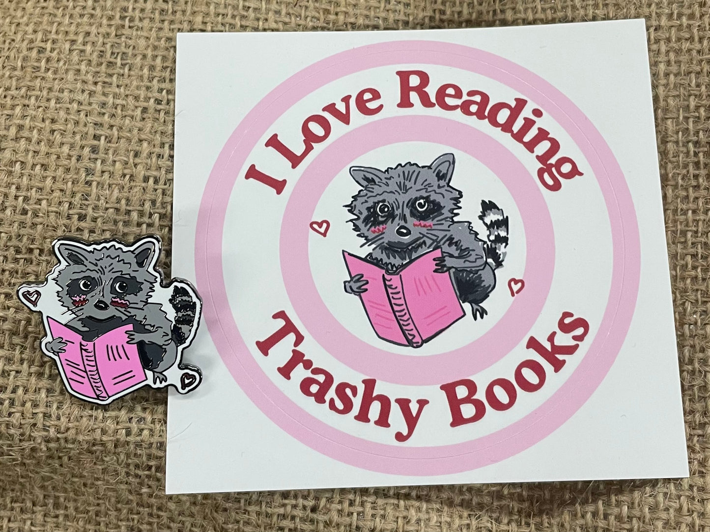raccoon enamel pin next to the i love reading trashy books sticker